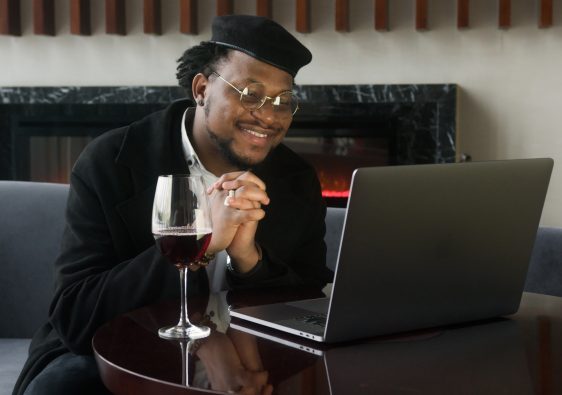 Handsome, black, plus size man smiling at laptop screen, sitting on couch, drinking wine